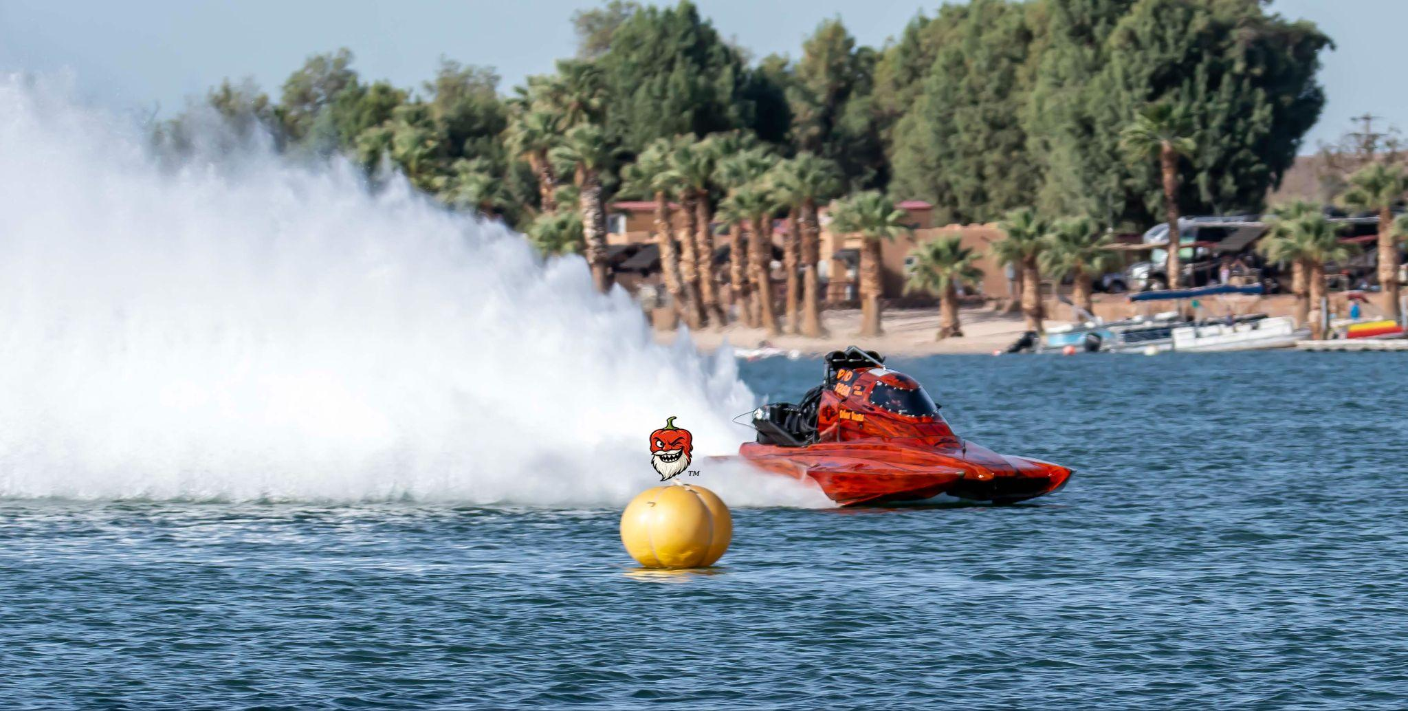 Red drag boat racing past a yellow buoy with a big white spray behind it, on a lake lined with palm trees