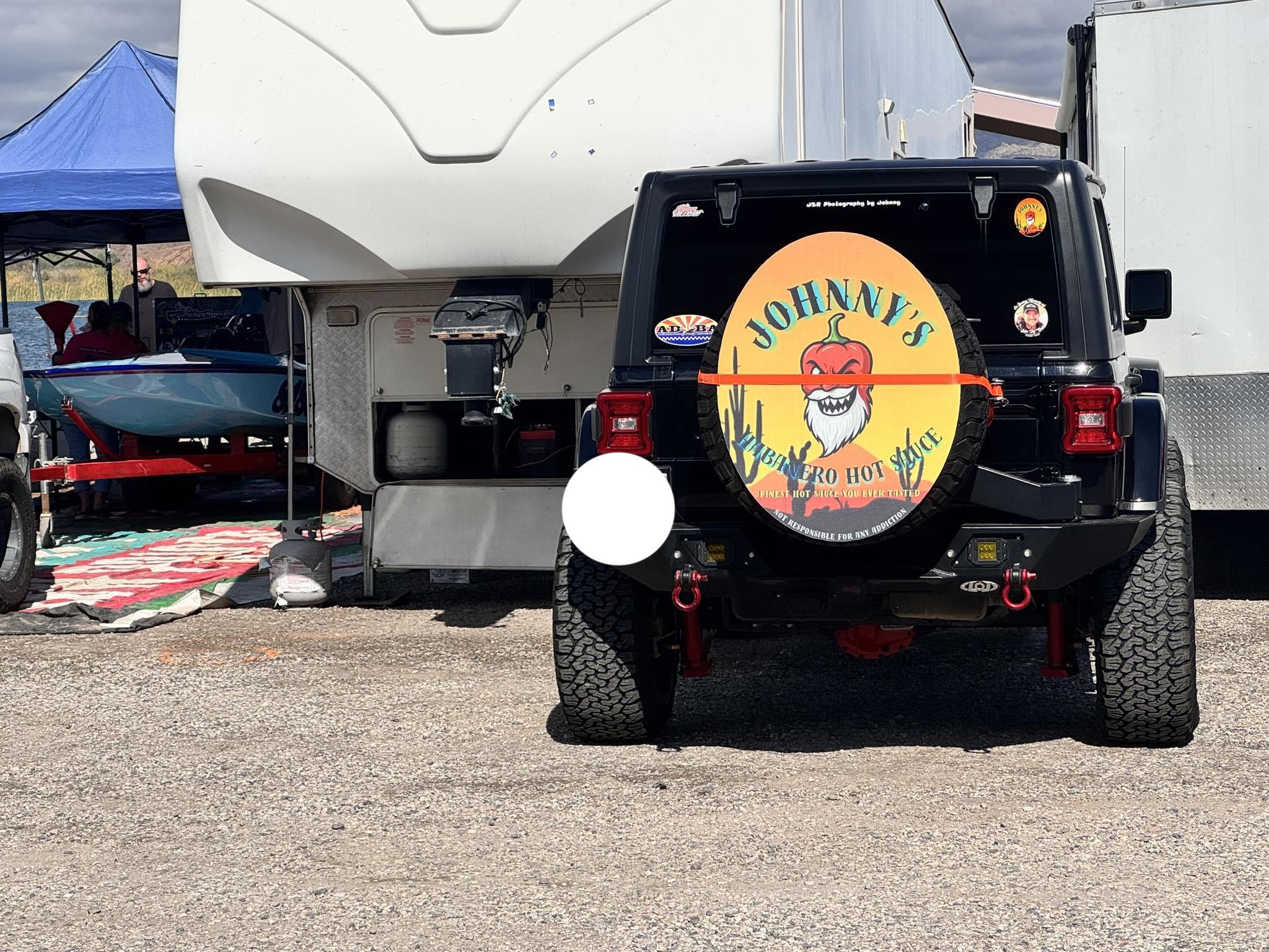 Rear view of a black Jeep with a Johnny's Habanero logo on its spare tire cover, parked at a campsite beside an RV and a boat